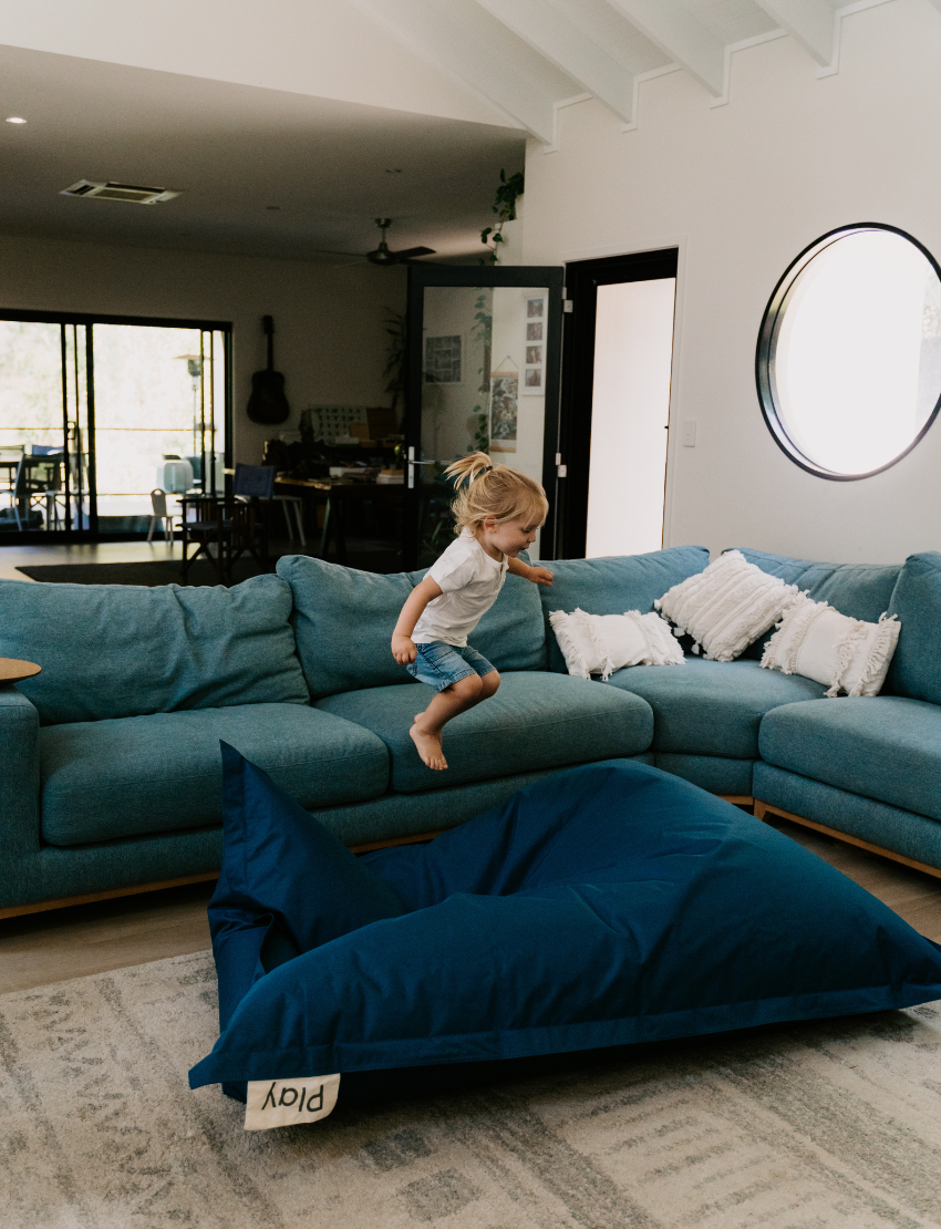 Two children immerse themselves in imaginative play on a large, blue Crash Mat by Sensory Play Australia, positioned in front of a gray sofa. Over the sofa's backrest hang colorful shirts, with one child sporting a vibrant tie-dye shirt and the other clad in a light blue shirt and shorts. Their activities encourage the development of motor skills through fun and creativity.