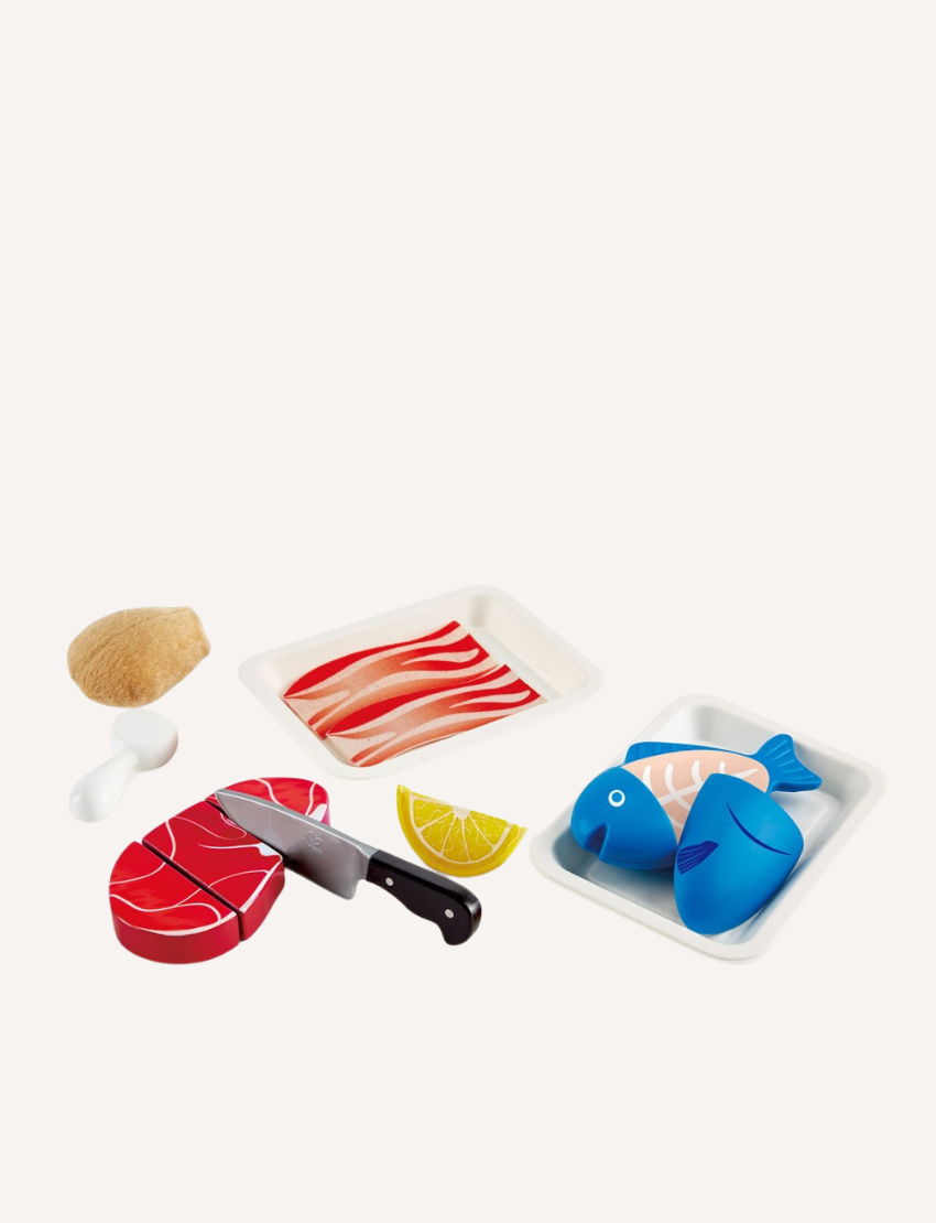 A display of Hape's Tasty Proteins toy set features a red meat cut, a blue fish, a knife, a lemon wedge, two sushi pieces, an oyster shell, and coconut halves. These items are arranged on white trays against a plain background.