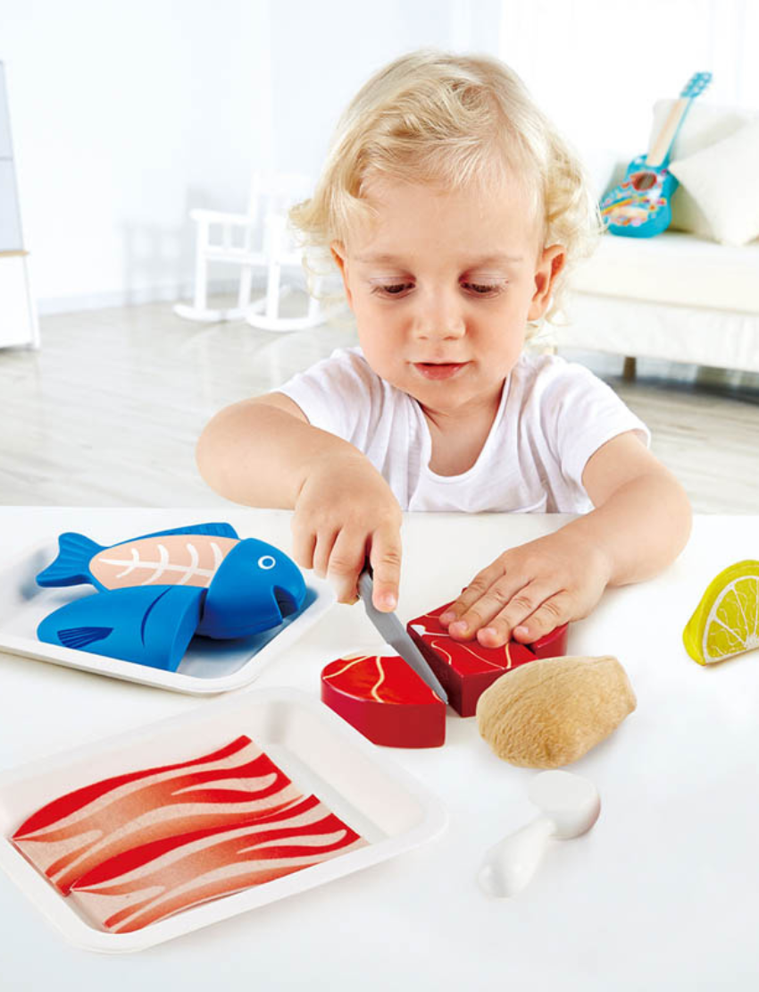 A display of Hape's Tasty Proteins toy set features a red meat cut, a blue fish, a knife, a lemon wedge, two sushi pieces, an oyster shell, and coconut halves. These items are arranged on white trays against a plain background.