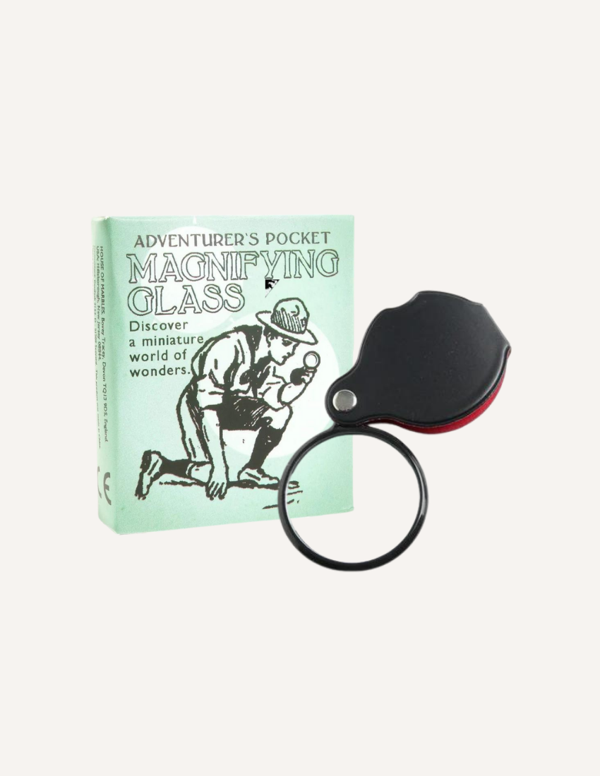 The image shows a small, boxed magnifying glass labeled "Adventurer's Magnifying Glass" from House of Marbles. The green box features an illustration of a person examining something with a magnifying glass. The actual magnifying glass is black with a foldable design.