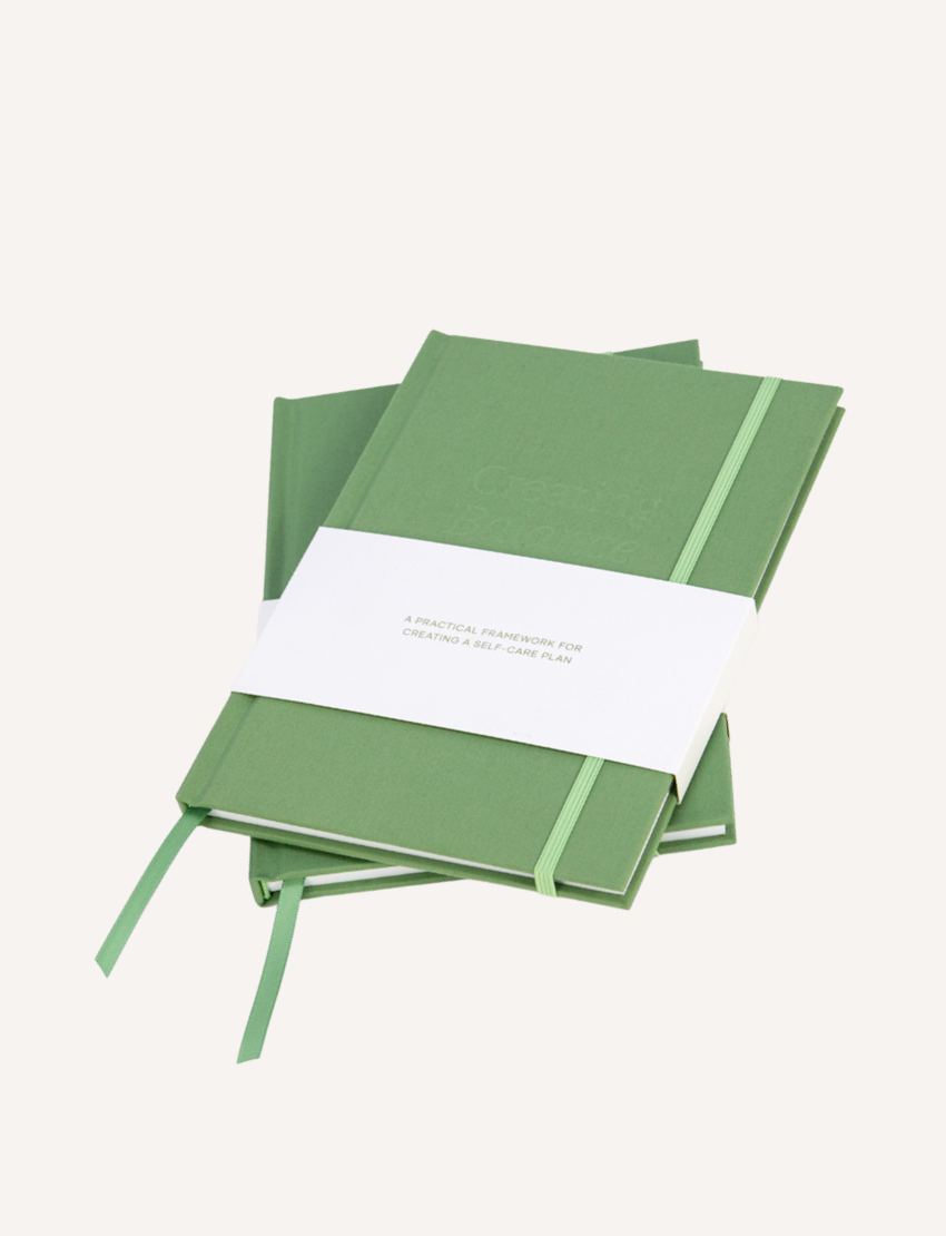 The Creating Balance Journal by Belucci is open on a white surface, displaying pages with printed forms and diagrams. Circular diagrams and bullet points appear on the right page, while a green ribbon bookmark marks the center.