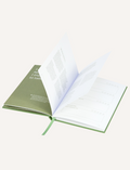 The Creating Balance Journal by Belucci is open on a white surface, displaying pages with printed forms and diagrams. Circular diagrams and bullet points appear on the right page, while a green ribbon bookmark marks the center.