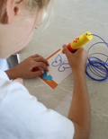 A vibrant Fine Motor Practice: Play Kit by The Play Way is open, revealing a delightful assortment of children's activity items. Included are ultra-light modeling clay, a 3D printing pen set designed to enhance fine motor skills, a gemstone excavation kit, and an array of small toys. The scene is set against a simple white background.