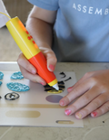 A vibrant Fine Motor Practice: Play Kit by The Play Way is open, revealing a delightful assortment of children's activity items. Included are ultra-light modeling clay, a 3D printing pen set designed to enhance fine motor skills, a gemstone excavation kit, and an array of small toys. The scene is set against a simple white background.