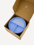 An open green and teal cardboard box from The Play Way's Tools For Eating - Bowls & Plates Kit contains a set of stackable silicone suction bowls, a set of stackable cups, and an upright blue divided baby plate. The items are neatly arranged, showcasing their self-feeding design.