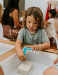 A vibrant Fine Motor Practice: Play Kit by The Play Way is open, revealing a delightful assortment of children's activity items. Included are ultra-light modeling clay, a 3D printing pen set designed to enhance fine motor skills, a gemstone excavation kit, and an array of small toys. The scene is set against a simple white background.