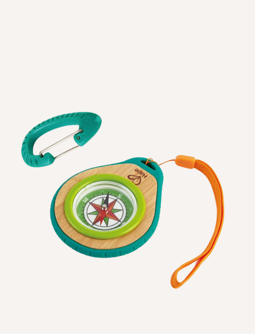 The Hape Compass Set includes a round compass featuring a red arrow, elegantly housed in a wooden base. It is encased in green and teal rubber with an attached orange wrist strap, and comes with a teal carabiner clip placed beside it.