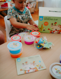 A vibrant Fine Motor Practice: Play Kit by The Play Way is open, revealing a delightful assortment of children's activity items. Included are ultra-light modeling clay, a 3D printing pen set designed to enhance fine motor skills, a gemstone excavation kit, and an array of small toys. The scene is set against a simple white background.