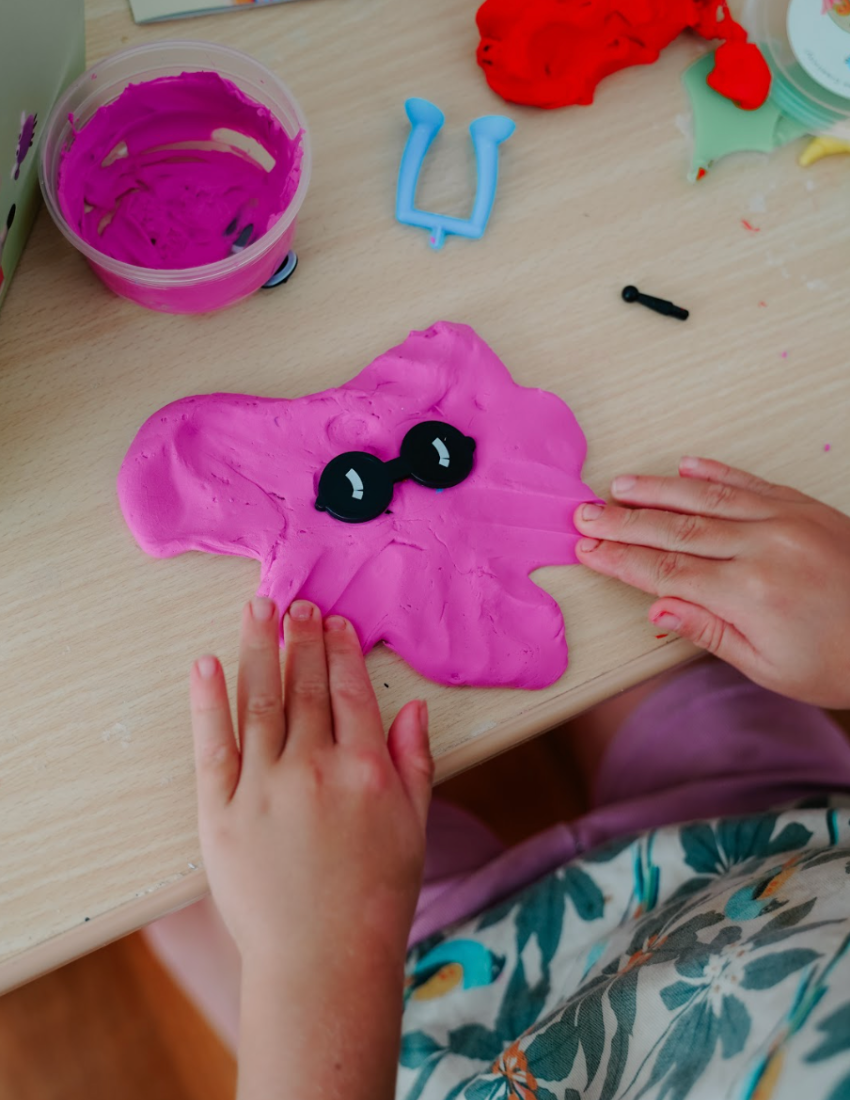 A vibrant Fine Motor Practice: Play Kit by The Play Way is open, revealing a delightful assortment of children's activity items. Included are ultra-light modeling clay, a 3D printing pen set designed to enhance fine motor skills, a gemstone excavation kit, and an array of small toys. The scene is set against a simple white background.