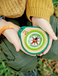 The Hape Compass Set includes a round compass featuring a red arrow, elegantly housed in a wooden base. It is encased in green and teal rubber with an attached orange wrist strap, and comes with a teal carabiner clip placed beside it.