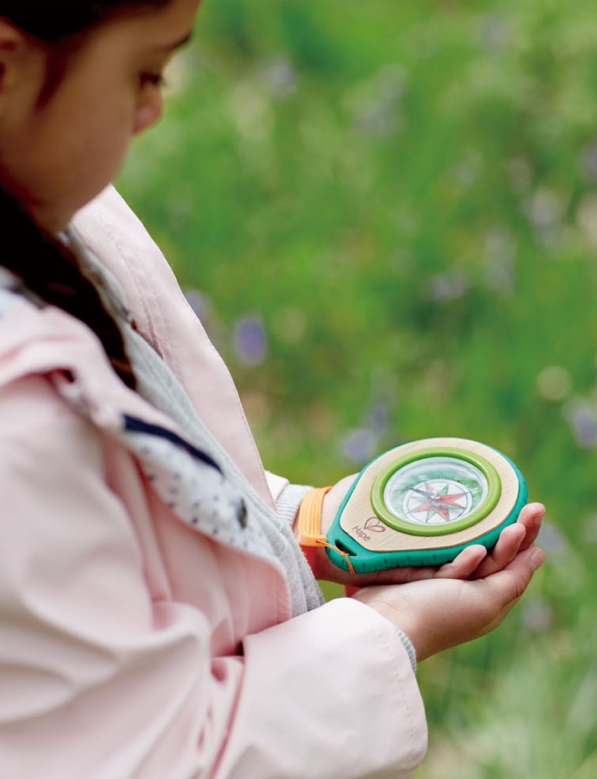 The Hape Compass Set includes a round compass featuring a red arrow, elegantly housed in a wooden base. It is encased in green and teal rubber with an attached orange wrist strap, and comes with a teal carabiner clip placed beside it.