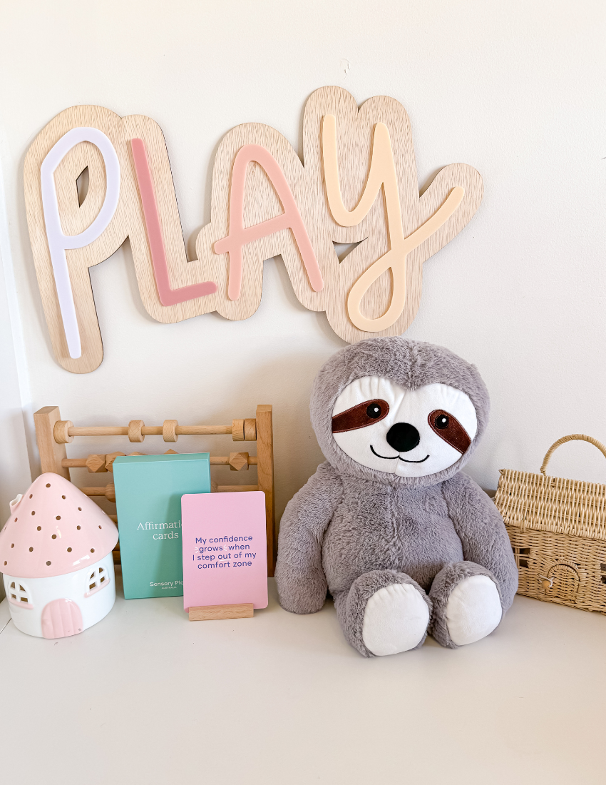 Introducing the Elizabeth Richards Weighted Sloth Toy: This adorable plush sloth showcases a cheerful expression, with distinctive brown and white facial markings. Its body is made of soft, light gray fabric complemented by white paws. Designed to sit upright against a plain off-white background, this comforting companion provides sensory regulation to promote emotional stability.