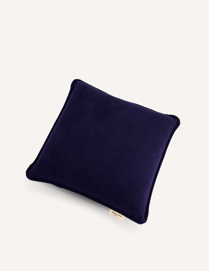 A Sensory Play Australia Vibrating Cushion, adorned in dark blue with a plain design, rests on a light-colored flat surface. The cushion features slightly rounded corners and includes a small fabric tag on one side.