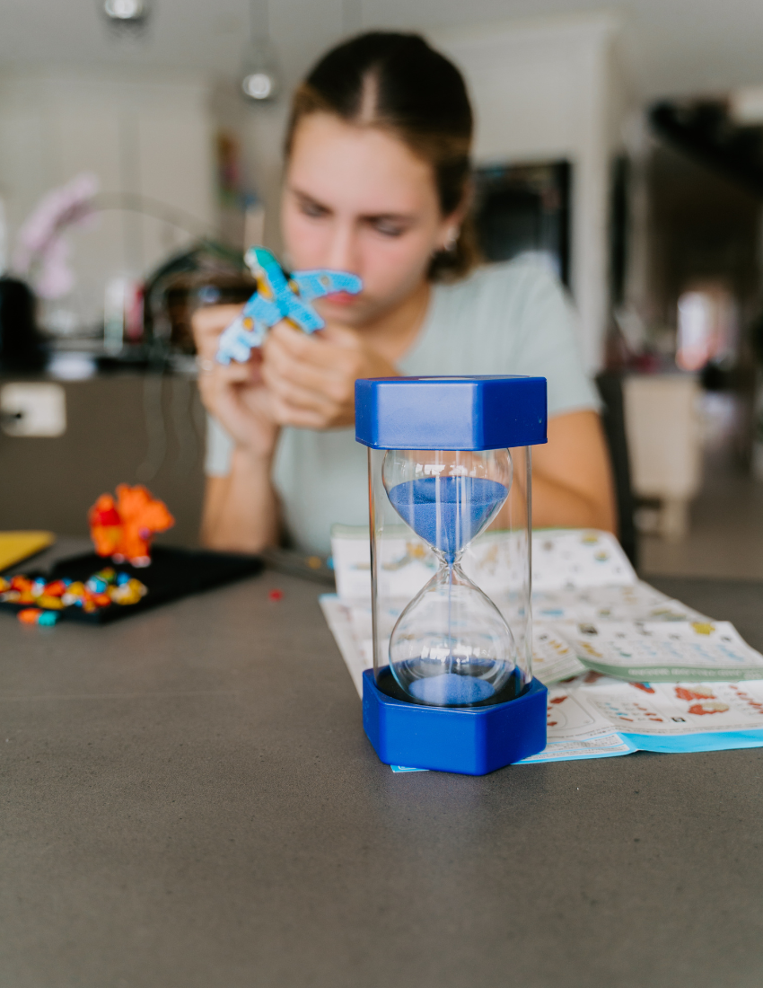 The Sand Timers Set by Sensory Play Australia, a collection of five vibrant sand timers ideal for managing time, is beautifully showcased. Each timer features hexagonal caps and bases in shades of purple, blue, green, orange, and black with coordinating sand inside. All the timers are neatly lined up against a plain white background.