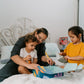 A parent with two kids is on a bed. They and one child are exploring The Play Way's Sleep Program box, while the other child holds a book. The bedroom features a decorative headboard and a patterned lamp.