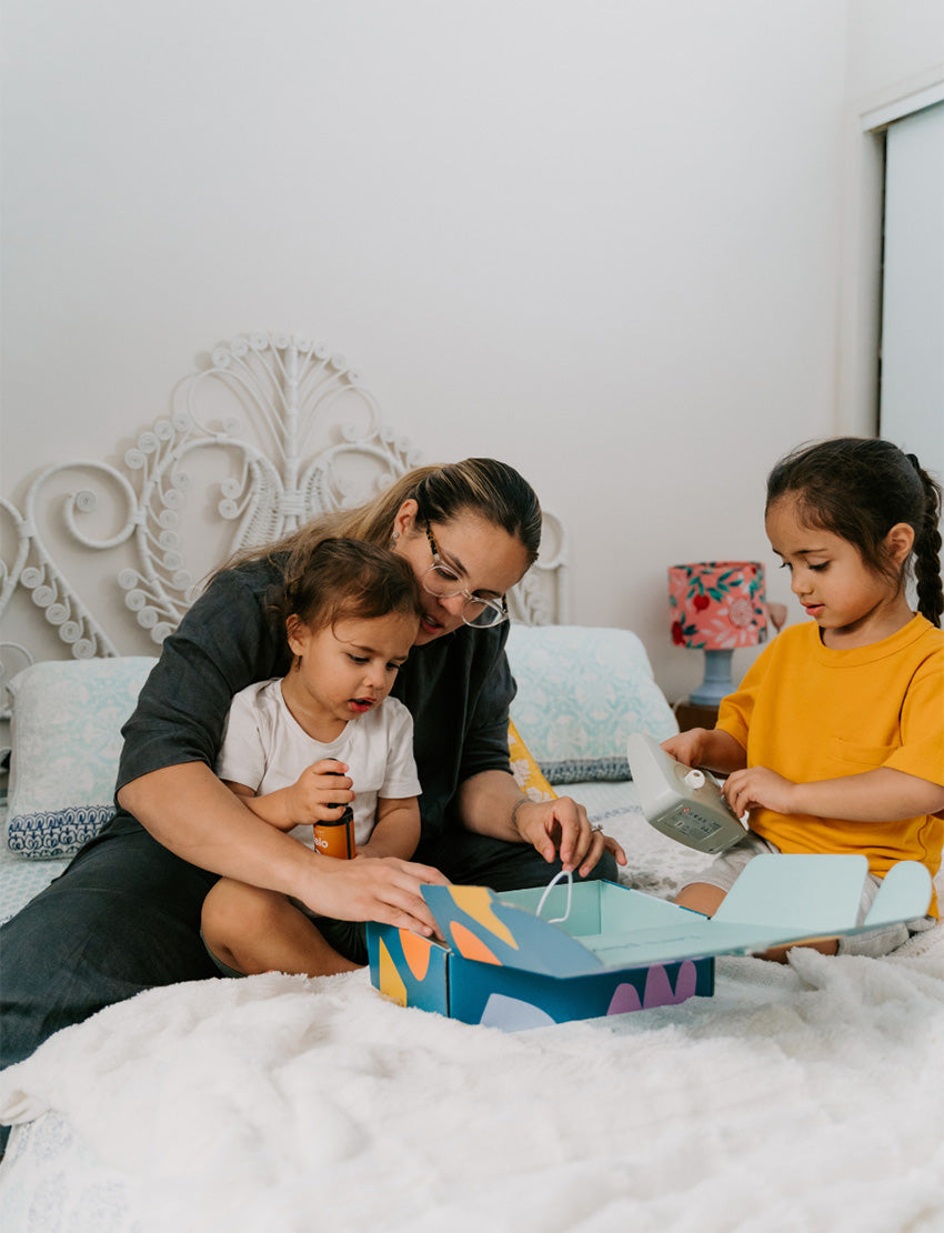 A parent with two kids is on a bed. They and one child are exploring The Play Way's Sleep Program box, while the other child holds a book. The bedroom features a decorative headboard and a patterned lamp.