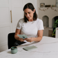 A woman in a white shirt sits at a marble table holding "Caregiver Wellbeing Training Program" by The Play Way, with another book in front of her. A green mug rests nearby, and a white cabinet and fireplace are visible in the background.