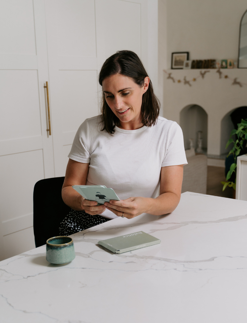 A woman in a white shirt sits at a marble table holding "Caregiver Wellbeing Training Program" by The Play Way, with another book in front of her. A green mug rests nearby, and a white cabinet and fireplace are visible in the background.