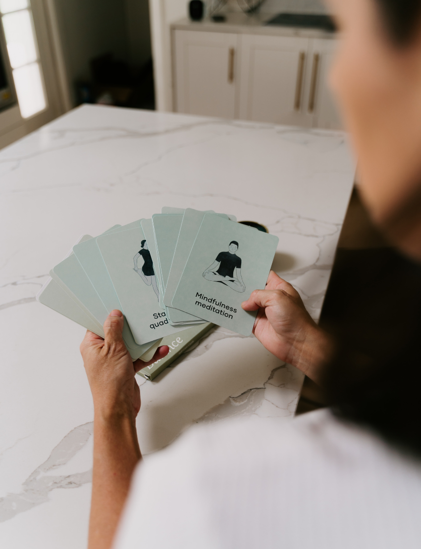 A woman in a white shirt sits at a marble table holding "Caregiver Wellbeing Training Program" by The Play Way, with another book in front of her. A green mug rests nearby, and a white cabinet and fireplace are visible in the background.