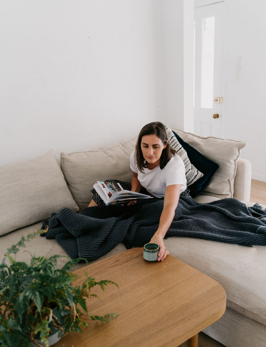 A woman in a white shirt sits at a marble table holding "Caregiver Wellbeing Training Program" by The Play Way, with another book in front of her. A green mug rests nearby, and a white cabinet and fireplace are visible in the background.