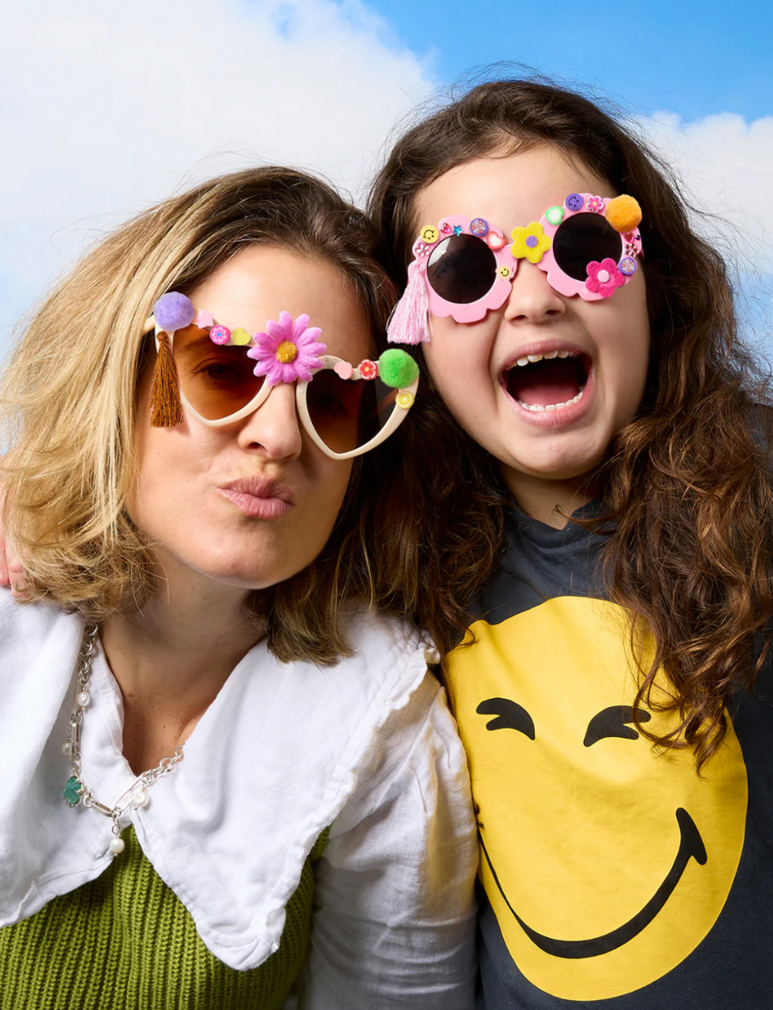 Two boxed craft kits feature a teal pair with heart-shaped sunglasses and a yellow pair with flower-shaped sunglasses. Both are labeled "Decorate Your Own Sunglasses" by Journey of Something and come with gem stickers for added flair.