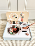 The Speedy Monkey Table Kitchen with 8 accessories is a children's wooden portable kitchen set that includes a stove complete with utensils, a pan, and pretend food. Made from FSC® certified wood, the design incorporates a backdrop featuring a clock and decorative elements. A booklet adorned with floral designs adds to this charming play area.