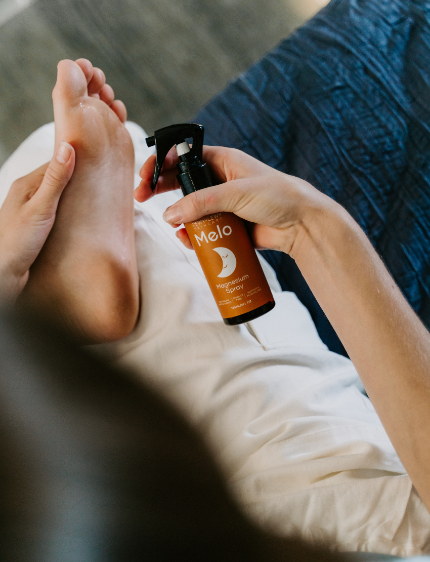 A brown bottle of Melo Magnesium Sleep Spray by Dewy + Lewy Skincare, featuring a black spray nozzle. The orange label is adorned with white text and a crescent moon illustration, highlighting its use for promoting restful sleep with calming essential oils. This product contains 125 mL (4.2 FL OZ).