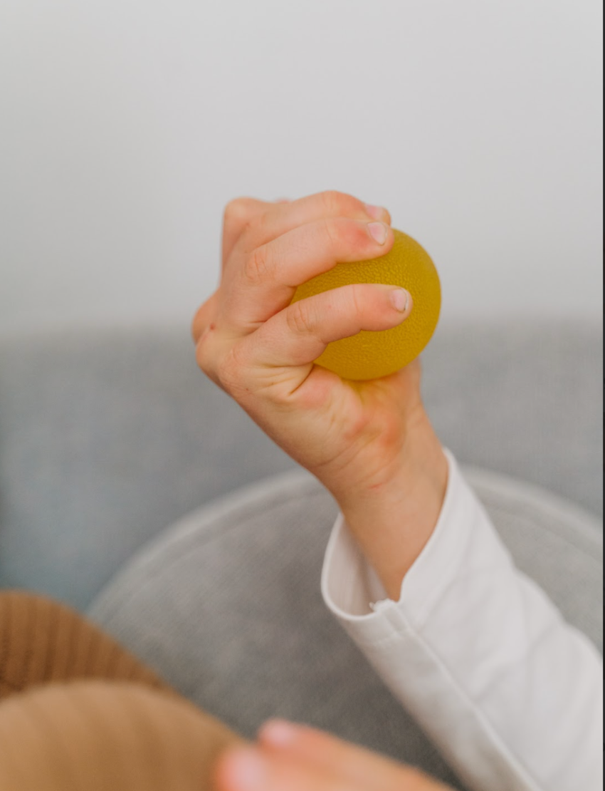 A teal box from The Play Way containing the Calm Story Kit is open, revealing a yellow squishy ball and a small booklet titled "Finding your calm." Both items are positioned visibly above the box. The box itself features a simple design with abstract shapes, ideal for sensory soothing and emotional regulation.