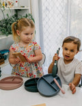 An open green and teal cardboard box from The Play Way's Tools For Eating - Bowls & Plates Kit contains a set of stackable silicone suction bowls, a set of stackable cups, and an upright blue divided baby plate. The items are neatly arranged, showcasing their self-feeding design.