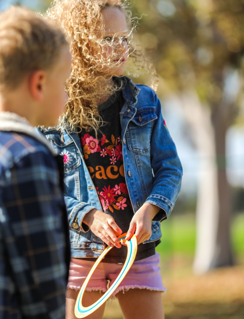 A freckled child with curly hair excitedly holds up an orange and blue Frisbee - Pack of 5 from The Play Way, in front of their face. Outdoors in a denim jacket, they smile amid the outdoor fun with blurred trees and grass adding a lively backdrop.