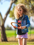 A freckled child with curly hair excitedly holds up an orange and blue Frisbee - Pack of 5 from The Play Way, in front of their face. Outdoors in a denim jacket, they smile amid the outdoor fun with blurred trees and grass adding a lively backdrop.