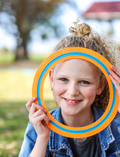 A freckled child with curly hair excitedly holds up an orange and blue Frisbee - Pack of 5 from The Play Way, in front of their face. Outdoors in a denim jacket, they smile amid the outdoor fun with blurred trees and grass adding a lively backdrop.