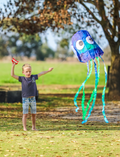 A young girl in a purple sweater and black pants holds a Jelly Kite by Tiger Tribe, designed as a blue jellyfish with big eyes and adorned with green and blue streamers. She is standing on a grassy field with trees in the background.