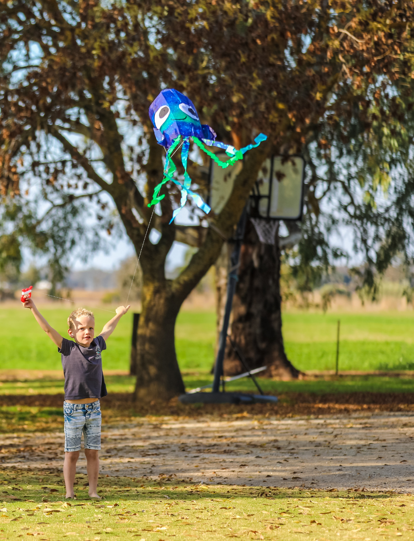 A young girl in a purple sweater and black pants holds a Jelly Kite by Tiger Tribe, designed as a blue jellyfish with big eyes and adorned with green and blue streamers. She is standing on a grassy field with trees in the background.