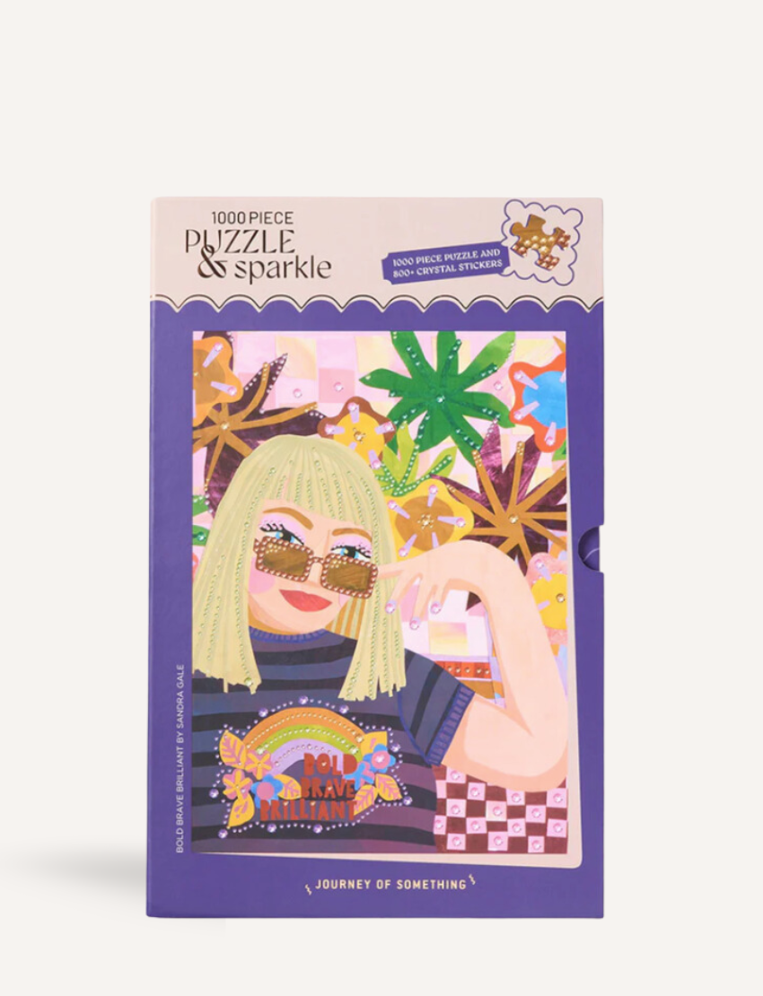 A 1000-piece puzzle box from Journey of Something titled "Bold, Brave, Brilliant" showcases a vibrant illustration featuring a person with blonde hair, sunglasses, and a rainbow shirt, adorned with palm trees, rainbows, and geometric patterns.