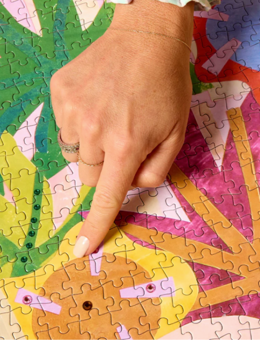 A 1000-piece puzzle box from Journey of Something titled "Bold, Brave, Brilliant" showcases a vibrant illustration featuring a person with blonde hair, sunglasses, and a rainbow shirt, adorned with palm trees, rainbows, and geometric patterns.