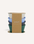 An open green and teal cardboard box from The Play Way's Tools For Eating - Bowls & Plates Kit contains a set of stackable silicone suction bowls, a set of stackable cups, and an upright blue divided baby plate. The items are neatly arranged, showcasing their self-feeding design.