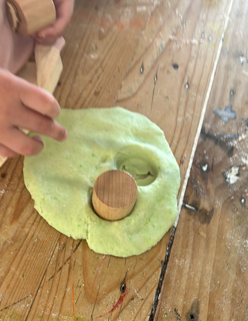 A box of the Fine Motor Practice: Playdough Kit by The Play Way, containing colorful playdough tubes and wooden stamps with various designs for creative expression, along with a booklet titled "Playdough Stamps." The box is adorned with abstract shapes and partially opened to reveal these sensory engagement tools, which are designed to enhance fine motor skills.