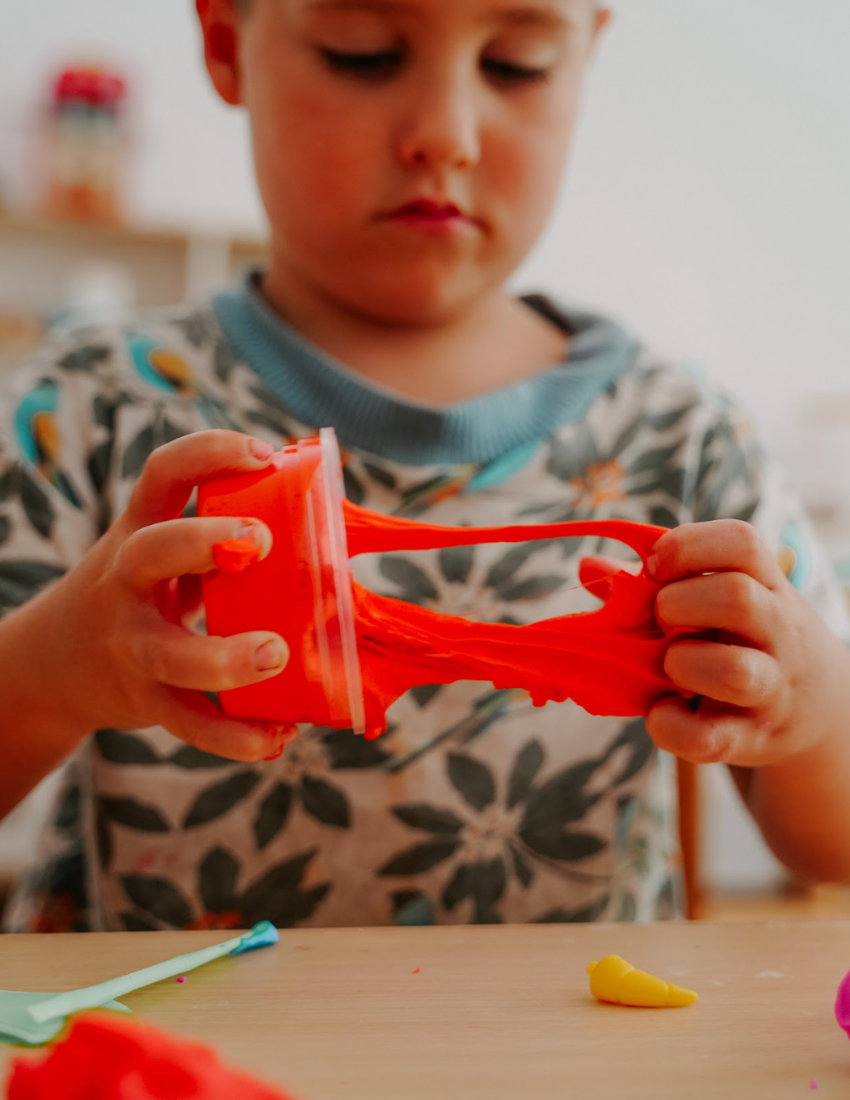 A box of the Fine Motor Practice: Playdough Kit by The Play Way, containing colorful playdough tubes and wooden stamps with various designs for creative expression, along with a booklet titled "Playdough Stamps." The box is adorned with abstract shapes and partially opened to reveal these sensory engagement tools, which are designed to enhance fine motor skills.