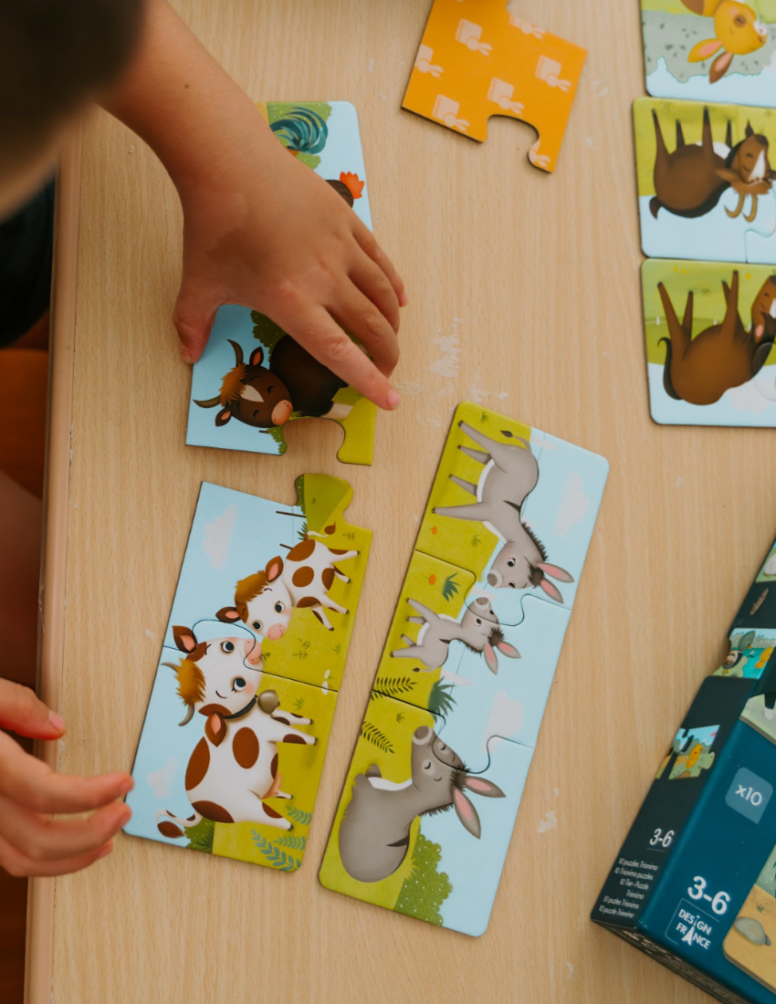 A large green box contains children's art supplies designed to foster creative expression: it features the Jar Melo Fine Motor Practice: Puzzle and Art Kit, which includes a pack of Giant Finger Paint Paper with a blue octopus and yellow crocodile, a Dot Painting Kit filled with colorful dots, and animal-themed tools that enhance fine motor skills.