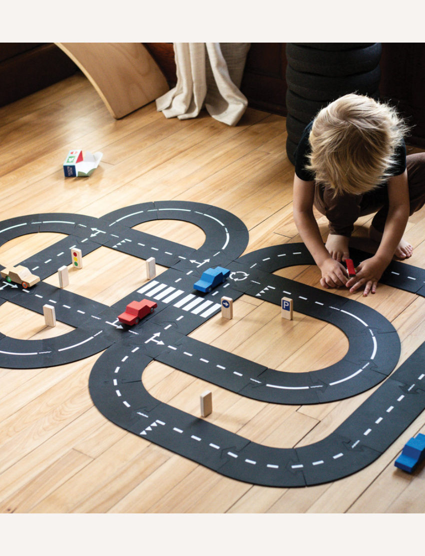 A child engages in play on a wooden floor with a black Waytoplay toy road set arranged in a figure-eight design. Various small colorful cars and road signs are positioned along the track. The child intently maneuvers the Waytoplay Back And Forth Car - Red.