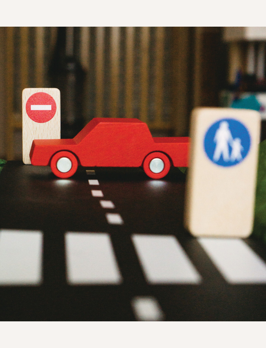 A child engages in play on a wooden floor with a black Waytoplay toy road set arranged in a figure-eight design. Various small colorful cars and road signs are positioned along the track. The child intently maneuvers the Waytoplay Back And Forth Car - Red.