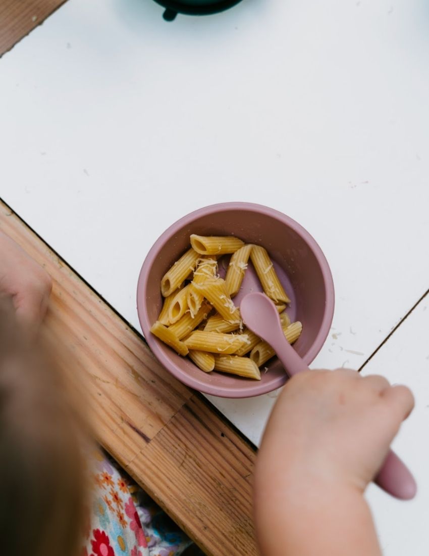 An open green and teal cardboard box from The Play Way's Tools For Eating - Bowls & Plates Kit contains a set of stackable silicone suction bowls, a set of stackable cups, and an upright blue divided baby plate. The items are neatly arranged, showcasing their self-feeding design.