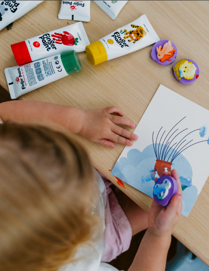 The Play Way's Fine-Motor Practice: Puzzle and Craft Kit is a large open green box designed to enhance sensory-rich creativity and early learning. It includes a variety of children's art supplies such as a coloring book, a finger paint set, colored shapes, markers, and activity cards, all showcased against a plain white background.