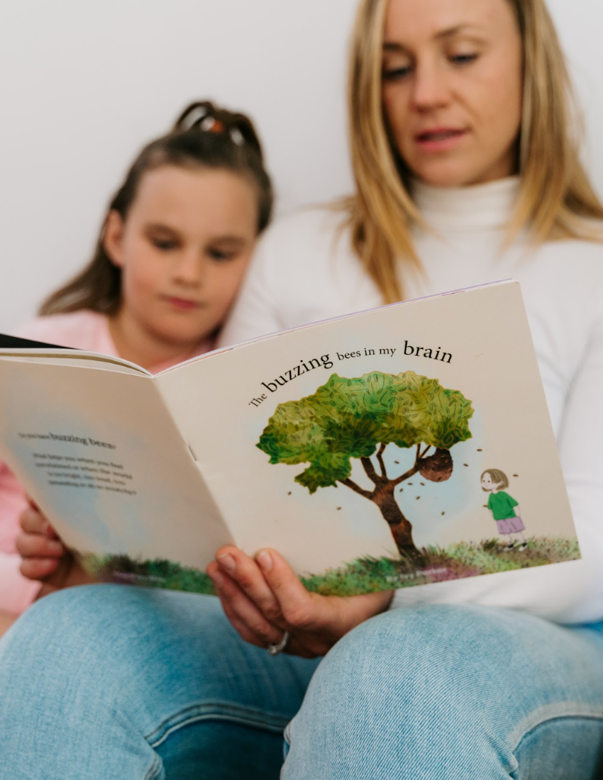 Cover of "The Buzzing Bees in My Brain Storybook" by Ivy Parker, a publication from The Play Way. The illustration features a girl standing near a tree with a beehive, encircled by bees. She is depicted wearing a green shirt, purple pants, and red shoes, embodying the sense of being overwhelmed amidst the buzzing chaos. This storybook is ideal for providing tips to parents on handling meltdowns.