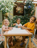 An open green and teal cardboard box from The Play Way's Tools For Eating - Bowls & Plates Kit contains a set of stackable silicone suction bowls, a set of stackable cups, and an upright blue divided baby plate. The items are neatly arranged, showcasing their self-feeding design.