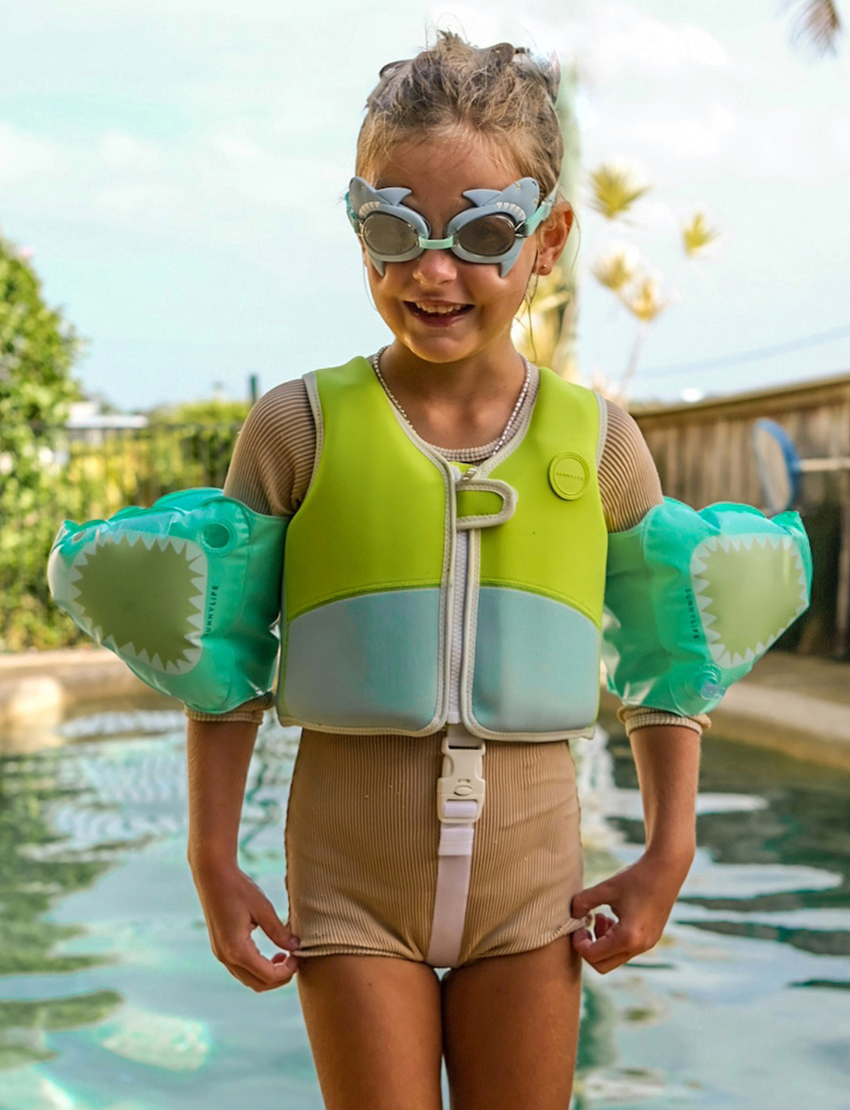 On a plain background, two pairs of Sunnylife's Mini Swim Goggles are displayed. The top pair is pink adorned with a shell design, while the bottom pair is blue with intricate shark details, including fins and teeth.