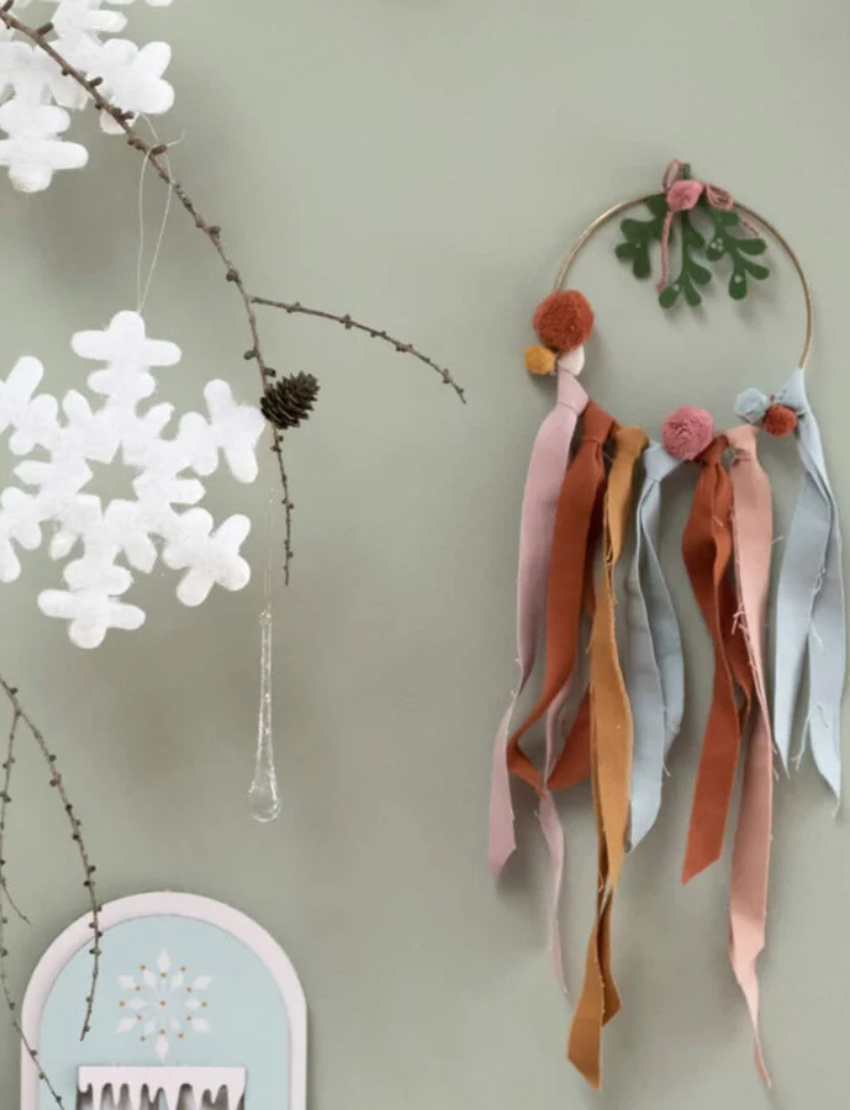 A Fabelab Christmas Wreath DIY kit featuring a decorative hoop with green leaves at the top, embellished with multicolored fabric ribbons and pom-poms in shades of pink, rust, and teal cascading from the bottom.