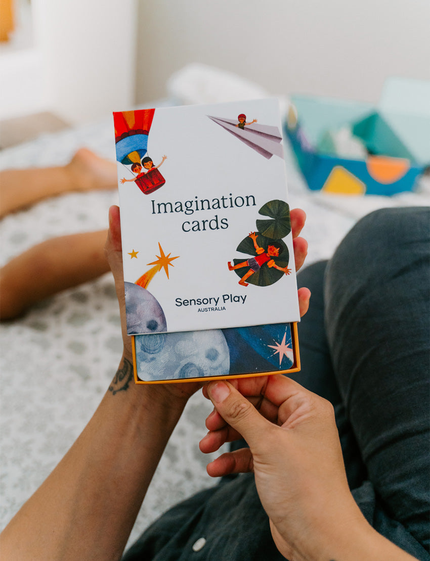 A parent with two kids is on a bed. They and one child are exploring The Play Way's Sleep Program box, while the other child holds a book. The bedroom features a decorative headboard and a patterned lamp.