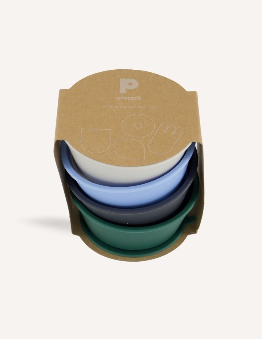 An open green and teal cardboard box from The Play Way's Tools For Eating - Bowls & Plates Kit contains a set of stackable silicone suction bowls, a set of stackable cups, and an upright blue divided baby plate. The items are neatly arranged, showcasing their self-feeding design.